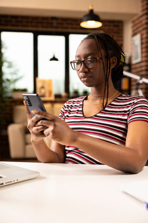 Black female freelancer in glasses, seated at desk with laptop, checking phone notifications and social media. African american remote manager chatting with colleagues on mobile device.の写真素材