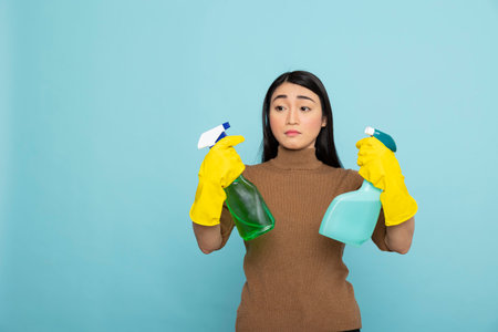 Pretty female janitor with yellow rubber gloves, holding two spray bottles and deciding on best disinfectant. Confident asian woman with liquid detergents ready for productive spring cleaning at home.の写真素材
