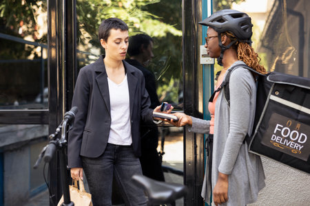 Black female worker accepts electronic payment from customer with a card on pos terminal. Courier ensuring transaction finalizes the purchase of fast food order from restaurant delivery.の写真素材