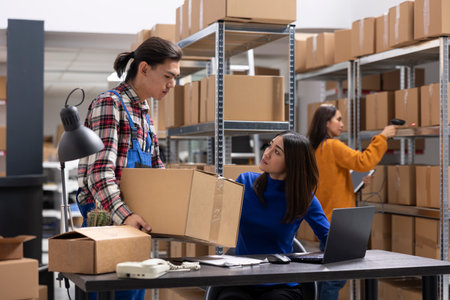 Asian workers team handles business logistics in a compact inventory room, taping boxes and attaching shipping labels. Small scale warehouse staff ensures retail product delivery.の写真素材
