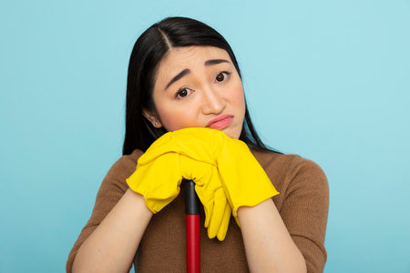 Close up of tired asian housemaid wearing yellow rubber gloves, resting head on hands. Female cleaner with unhappy expression takes break, feeling overwhelmed by messy household chores.の写真素材