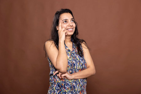 Empowered indian woman posing with her finger on forehead, embodying thoughtfulness and determination. Smiling female model looking upwards, reflecting on a positive memory in brown background studio.の写真素材