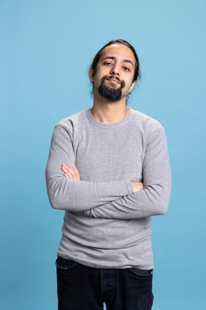 Young adult in casual wear standing with his arms crossed on camera, posing with confidence against a blue background. Man with a beard and long hair, calm natural attitude.の写真素材