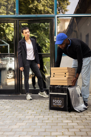 Black delivery worker passes stack of hot pizzas to client at the doorstep, restaurant takeaway service. Fast food large cardboard boxes order delivered directly to a woman customer.の写真素材