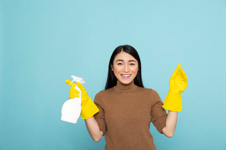 Smiling asian housekeeper holding cleaning spray in one hand while gesturing above isolated...