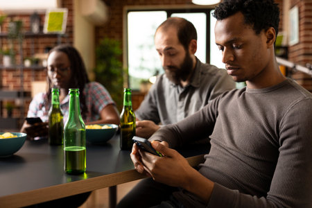 Close up of african american man using smartphone, with bottle of beer nearby on table. Black male individual engages in online browsing while sitting with friends in brick walled room.の写真素材