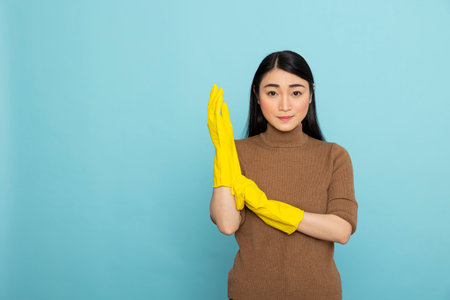 Asian woman putting on bright yellow rubber gloves, standing confidently and preparing to start cleaning. Portrait of housekeeper ready for household chores over isolated background.の写真素材