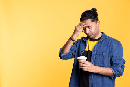 Tired indian man sipping fresh coffee from disposable paper cup early in the morning to wake up and be energized. Person drinking hot beverage from recycled takeaway cup, studio background.の写真素材