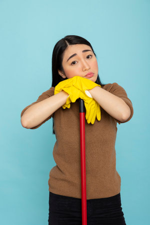 Portrait of tired asian female cleaner leaning on broomstick with sad expression. Young housekeeper wears yellow gloves, reflecting on overwhelming chores after long, exhausting day of cleaning.の写真素材