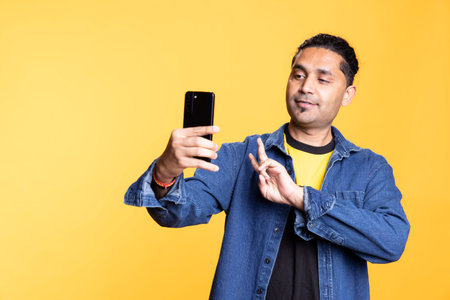 Indian young person using his mobile phone to take selfies from all angles. Male social media user taking pictures using his smartphone camera, smiling happily against studio background.の写真素材