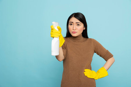 Female housekeeper confidently holding white spray bottle against isolated background. Focused janitor with hand on waist and grasping liquid detergent, ready to clean with precision.の写真素材