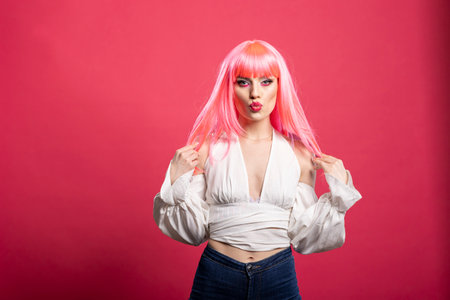 Stylish female adult in fashionable blouse and pink wig pouting confidently while holding hair. Glamorous caucasian woman standing against isolated backdrop with flirty expression.の写真素材