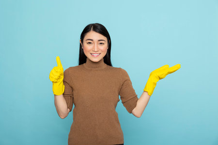 Happy housemaid wearing cleaning gloves points to right with open hand and gives thumbs up. Cheerful female janitor gestures to blank copy space on blue background for promotional ads.の写真素材
