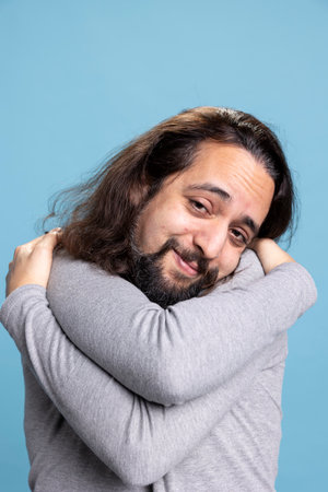 Young man practicing self love and self appreciation by hugging himself, being satisfied and fulfilled with his ego in studio. Powerful relaxed person shows self esteem and self care on camera.の写真素材