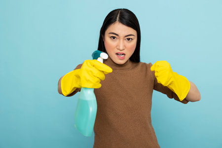 Annoyed asian housemaid in rubber gloves holding detergent spray in aggressive pose. Angry female janitor standing in fighting stance, shows frustration while dealing with tough house chores.の写真素材