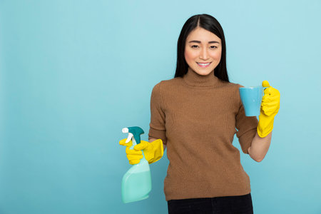 Cheerful cleaner takes break from chores with coffee in hand and cleaning product, standing joyfully in blue background studio. Housemaid stands with mug and spray bottle, resting from household tasksの写真素材