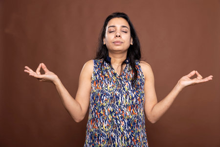 Beautiful woman with closed eyes and arms outstretched, standing in meditation against brown background. Portrait of Indian female individual immersed in a peaceful meditation session.の写真素材