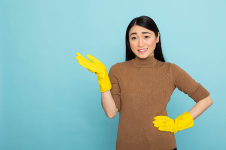 Asian housekeeper wearing rubber gloves, posing with one hand on waist and other gesturing to left. Young female janitor stands against isolated background, presenting cleaning service and products.の写真素材