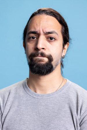 Displeased disappointed young man posing with arms crossed in studio, acting serious in casual wear against blue background. Young person with long curly hair and a beard on camera.の写真素材