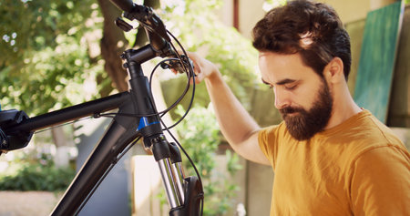 Sports-loving male loosening and removing damaged wheel from bike front fork with expert multitool outside. Committed man examining bicycle handlebar stem and headset in yard. Dolly zoom-in.の写真素材