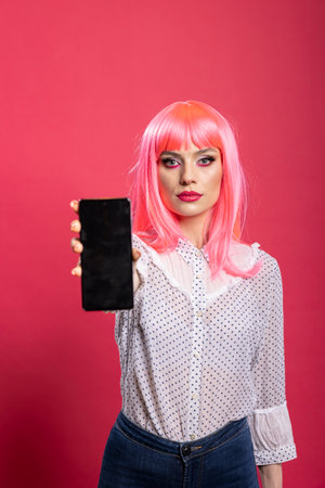 Casual young woman with vibrant pink hair presenting cellphone with empty screen, ideal for copy space or app preview. Caucasian female holding mobile device toward camera, displaying black display.の写真素材