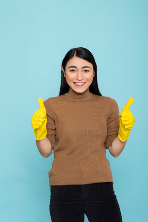 Happy asian housekeeper posing in blue background studio, expressing satisfaction with household chores. Young female janitor gesturing two thumbs up during productive morning cleanup routine.の写真素材