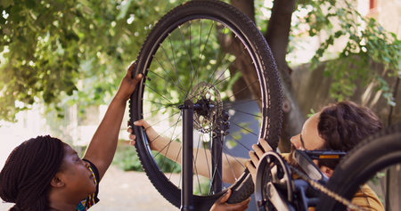 African american girlfriend carrying wheel to reattach on bicycle body with caucasian boyfriend. Sports-loving multiethnic couple carefully securing dismantled bike tire outside.の写真素材