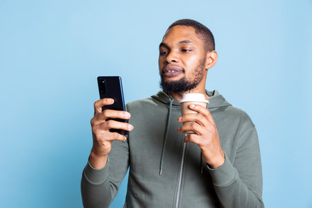 Positive friendly guy drinks coffee and checks his smartphone, answering to online messages being cheerful against blue background. Happy man celebrating good news and messaging.の写真素材