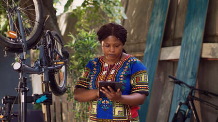 Detailed view of sports-loving black woman searching on phone tablet for bike summer maintainance. African american female grasping smart device while inspecting and maintaining bicycle.の写真素材