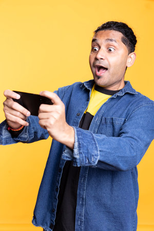 Happy joyous indian man playing online video games against studio background, single player enjoying a competition tournament on mobile phone. Young adult with a cheerful expression.の写真素材