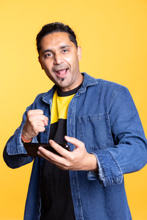 Happy gamer celebrating after winning game on gaming console, standing against studio background. Delighted Indian man bragging after being victorious in video gaming competition, defeating enemies.の写真素材