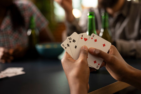 Closeup of african american hand gripping playing cards during fun indoor activity with friends, surrounded by snacks and refreshments. Black person holding pair of cards during casual gaming session.の写真素材