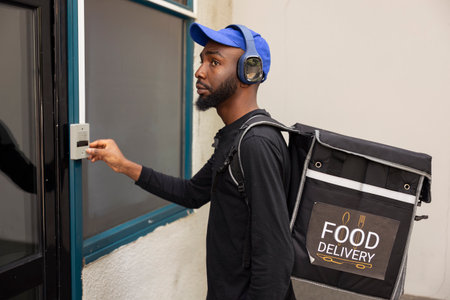 African american worker approaches the doorway to drop off a hot meal order, ringing the intercom to deliver the food. Young adult employee ensuring an express delivery takeaway service.の写真素材
