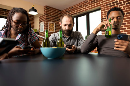 Male and female individuals focus on cellphones, browsing social media while sitting together at table with drinks and snacks. Diverse group of friends hanging out, engaged with mobile devices.の写真素材