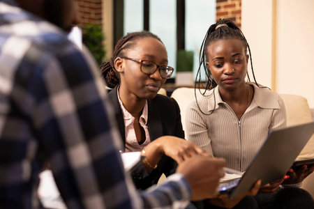 African American woman wearing glasses, using laptop to give business project updates to her colleagues. Female coworkers seated in office couch, discussing marketing strategies and solutions.の写真素材
