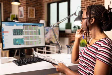 Black woman holds documents and pen, analyzing data on computer and updating business metrics from home. Female freelancer analyzing charts on screen while referencing notes from clipboard.の写真素材