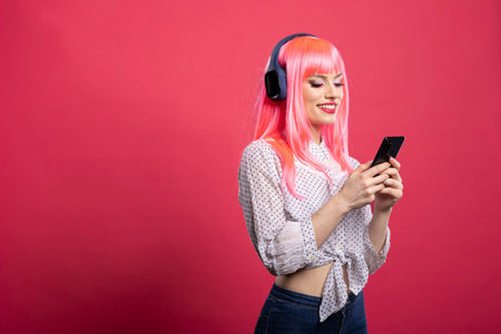 Joyful female individual with wireless headset, watching hilarious video on her smartphone. Happy white woman wearing pink wig, enjoying music and looking at mobile device, browsing on social media.の写真素材