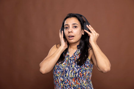 Beautiful indian woman in headphones enjoying favorite playlist. Relaxed female individual touching her wireless headset while looking at camera, standing against isolated background.の写真素材