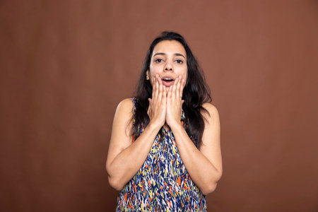 Stunned Indian woman with hands on cheeks and eyes wide open, expressing dramatic disbelief. Beautiful female model stands against brown background, looking at camera in shock and amazement.の写真素材