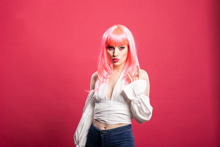 Beautiful caucasian woman with pink hair and pursed lips, flashing playful rock hand gesture. Fashionable young lady expresses bold personality against pink background in energetic studio portrait.の写真素材