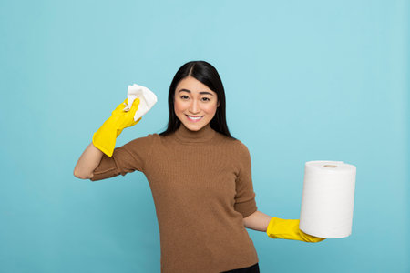 Asian housekeeper wearing rubber gloves holding paper towel, standing ready for daily chores. Portrait of female janitor posing with roll of kitchen tissue to ensure hygiene and cleanliness.の写真素材