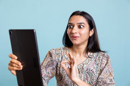Confident upbeat girl sharing a story with best friend over web video call, standing against blue background with smartphone. Happy woman talking with mate online on teleconference.の写真素材