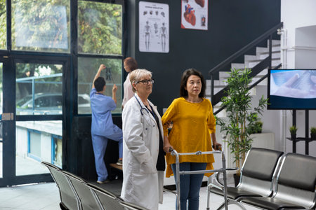 Doctor assisting patient while she uses walking frame during rehabilitation therapy, having a checkup appointment. Physical therapy discussion with mobility issues, healing process.の写真素材