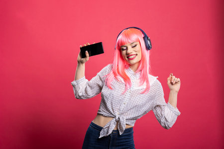 Excited woman with pink hair and headphones, enjoying music through cellphone. Cheerful young lady in casual outfit, dancing and having great time in studio, standing against isolated background.の写真素材