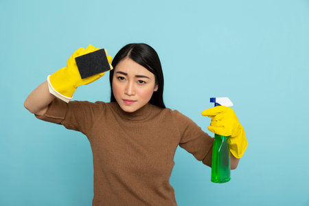 Asian housemaid with focused expression holds spray bottle and sponge, ready to tackle household chores. Confident female janitor posing with cleaning supplies, prepared to maintain spotless surfaces.の写真素材