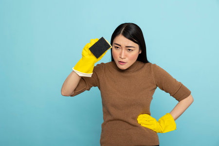 Portrait of serious asian woman in yellow rubber gloves, holding cleaning sponge and hand on waist, staring ahead with intent. Housemaid standing ready for chores with confidence and precision.の写真素材