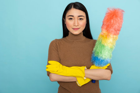 Portrait of confident female janitor wearing yellow rubber gloves and holding feather duster, ready for cleaning duties. Asian housekeeper standing with crossed arms against blue background.の写真素材