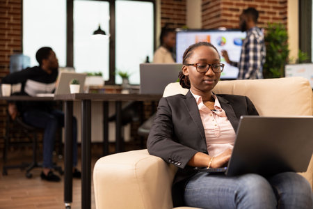 Black female company advisor using laptop, seated on couch, analyzing marketing strategy and compiling reports. African american woman manager comfortably working on business project in startup officeの写真素材