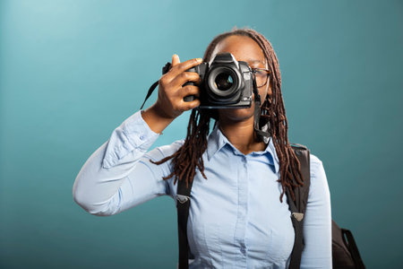 Portrait of black female photographer holding her DSLR, ready for client shoot, showcasing her professionalism. African american woman standing with camera, taking a shot in modern studio.の写真素材