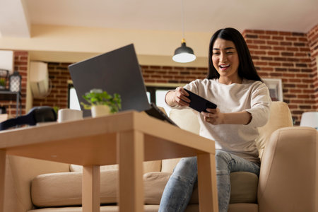 Excited remote female manager taking pause on work, playing video games on cellphone. Cheerful businesswoman resting and enjoying mobile gaming entertainment in brick wall apartment.の写真素材